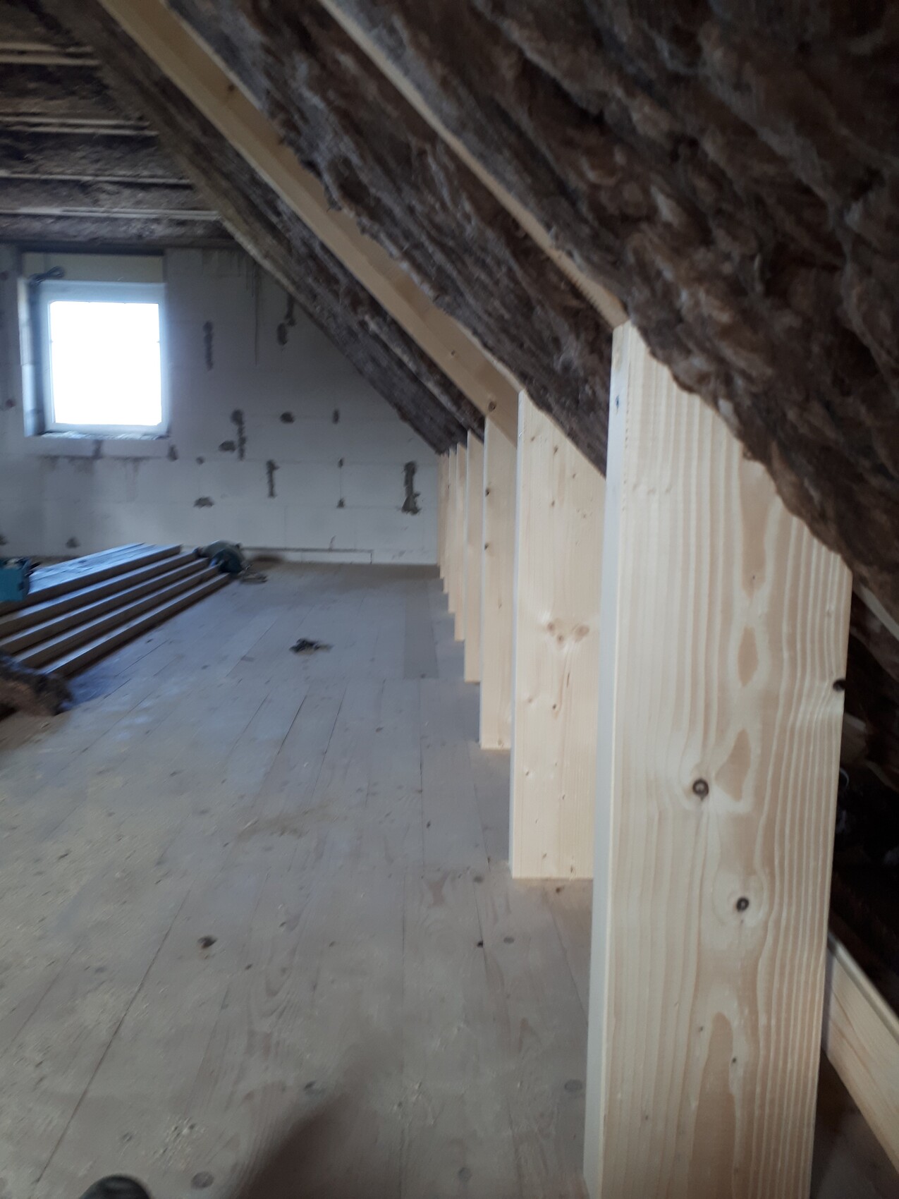 Dachboden-Baustelle: sichtbare Holzpfosten, Dämmung am Dach, Holzboden, Fenster am Ende