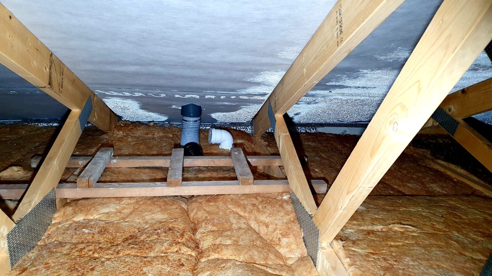 Dachboden mit Holzrahmen, orange Isolierung und graue/weiße Rohre.