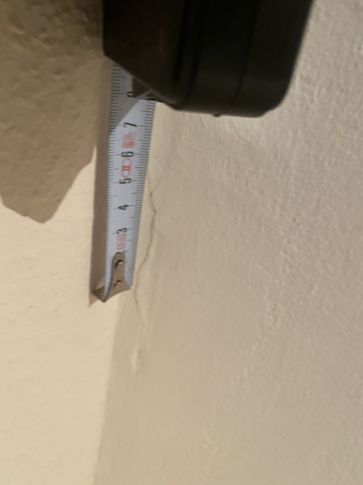 Maßband in Halterung an einer hellbeigen Wand; Messskala mit Zahlen ist sichtbar.