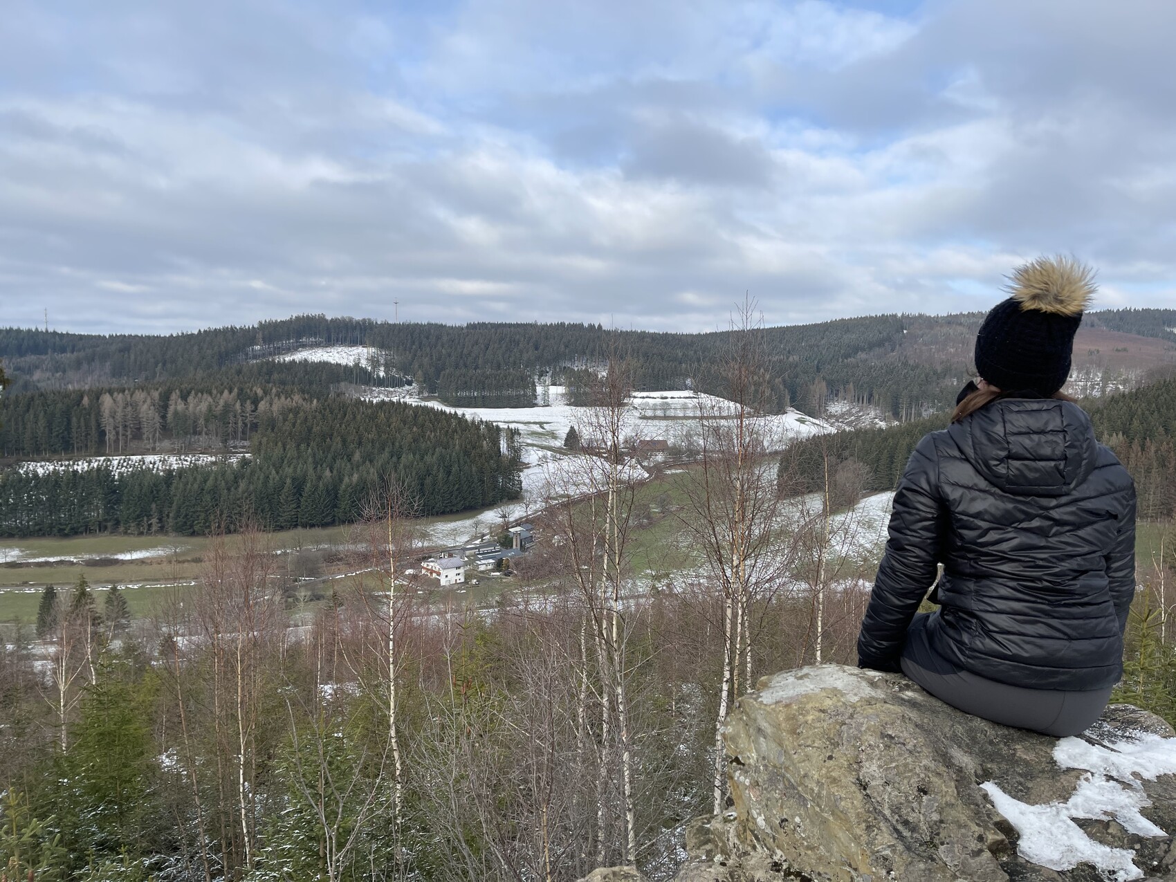 Person sitzt auf Felsen und blickt über verschneite Wald- und Wiesenlandschaft mit Häusern.