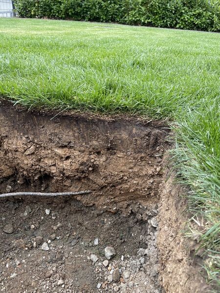 Grasfläche über freigelegtem Erdgraben mit sichtbarer Rohrleitung.