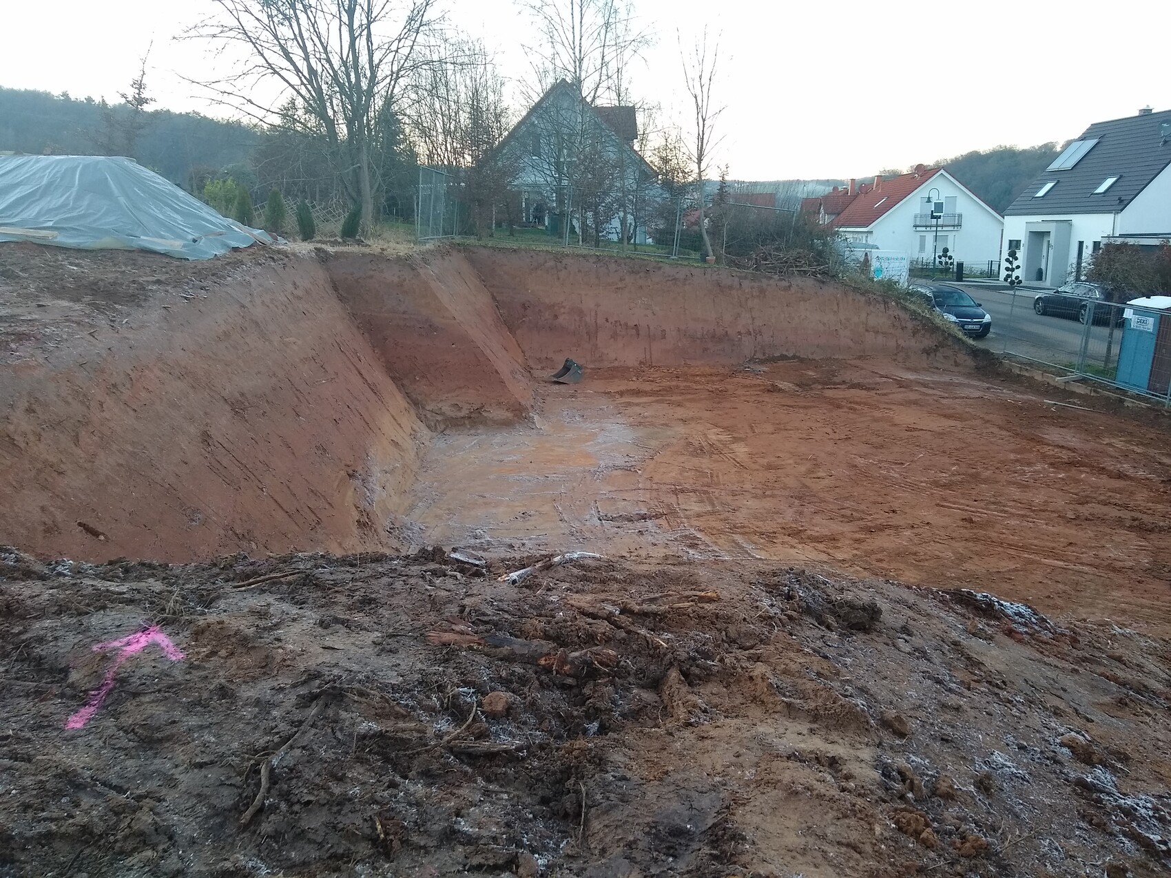 Große Baugrube mit rotem Erdreich; geneigte Wände, links eine Abdeckung, Häuser im Hintergrund.