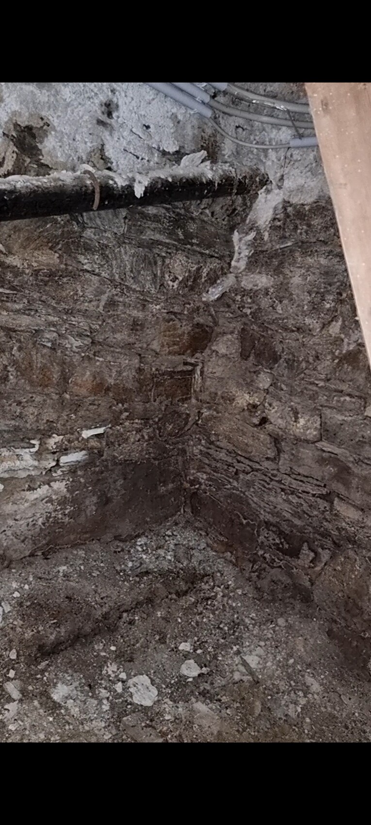 Ausgehobene Baugrube im Keller mit rauer Steinwand, Staub und sichtbaren Rohren.
