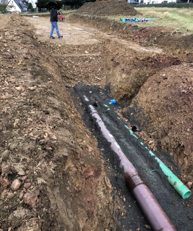 Baustelle mit ausgehobenen Gräben, Rohren und großen Erdehügeln; Arbeiter im Hintergrund.
