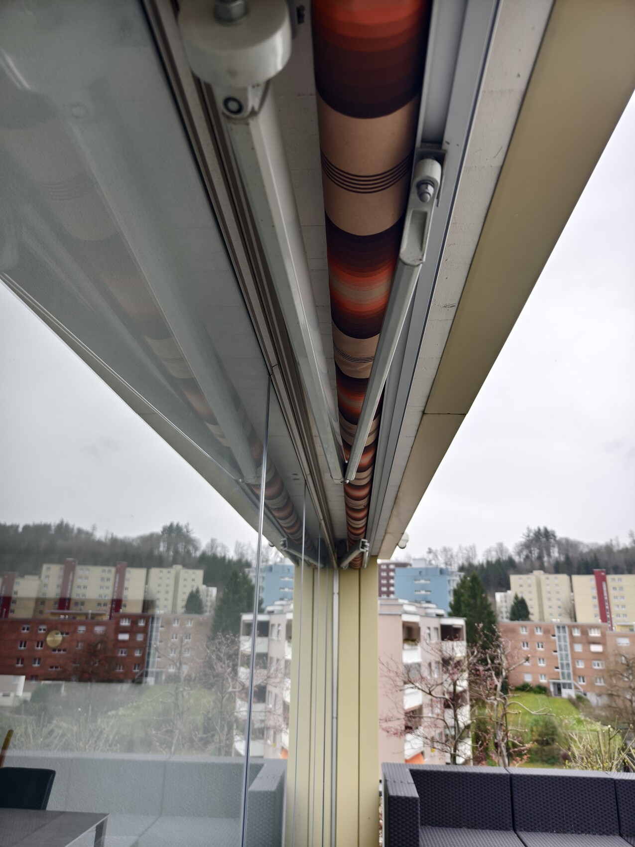 Balkon-Sonnenstore von unten mit rot-braunen Rollen; Stadtblock im Hintergrund.