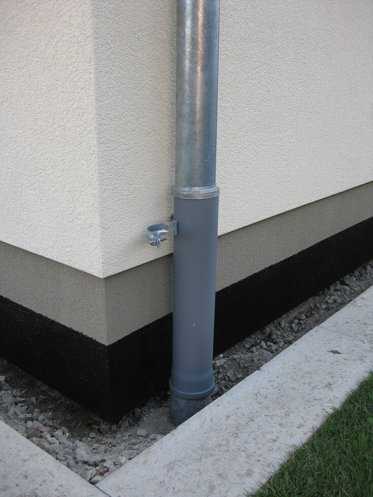 Außenwand eines Hauses mit vertikalem Metall-Downpipe am Fundament neben Kies und Gras