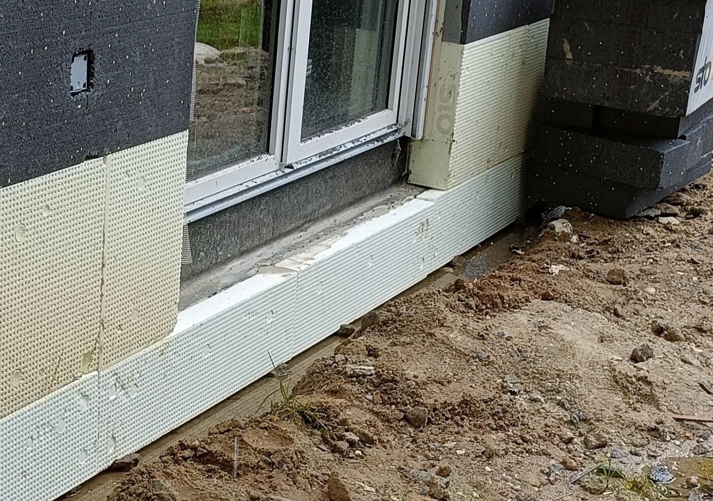 Außenwand eines Hauses im Bau mit Dämmplatten, Fenster und Erdreich am Fundament.