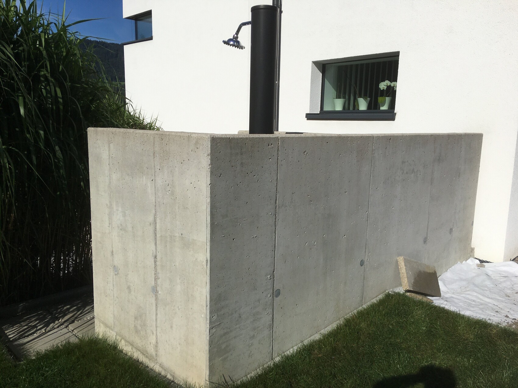 Große graue Betonwand vor weißem Haus mit Fenster und Topfpflanzen; Außendusche an schwarzem Rohr.
