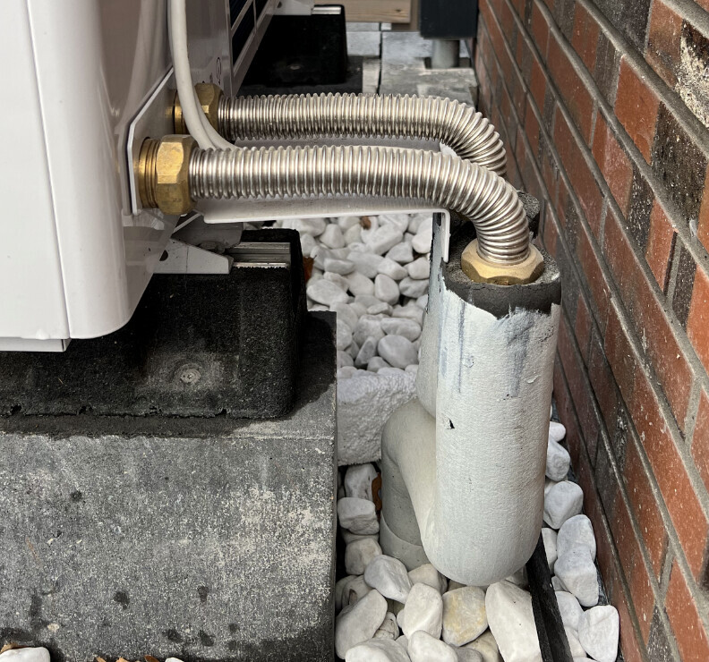 Außengerät einer Klimaanlage mit flexiblen Metallleitungen, montiert auf Beton neben einer Ziegelwand.