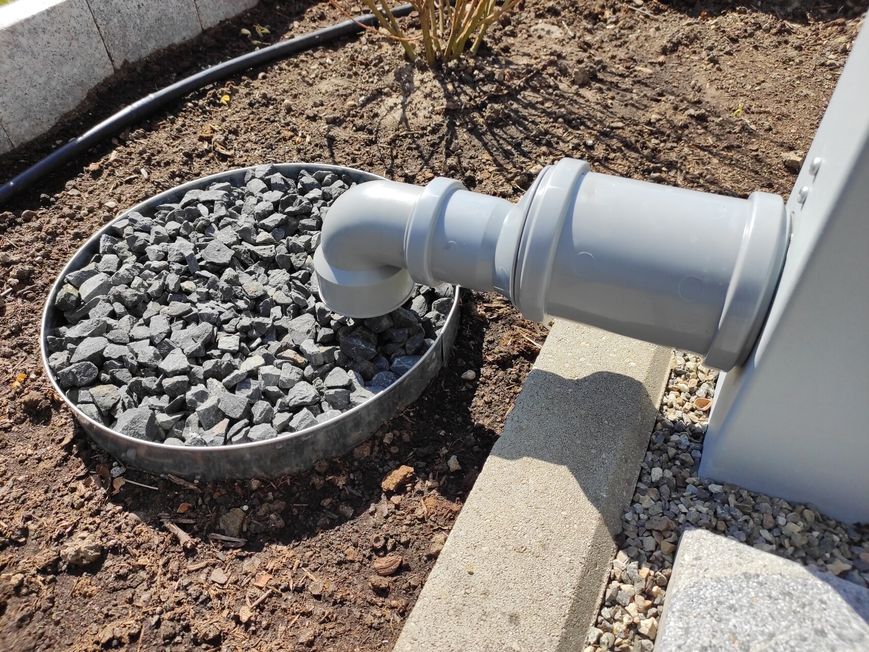 Graues PVC-Abflussrohr mit 90°-Bogen führt in eine runde Metall-Schale mit Kies im Außenbereich.