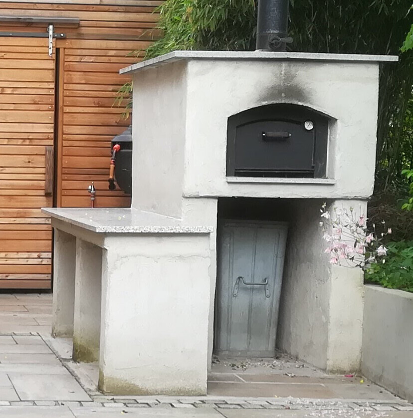 Außengrill/Backofen aus Beton mit oberem Backofenfach und darunterem Stauraum, Gartenumgebung.