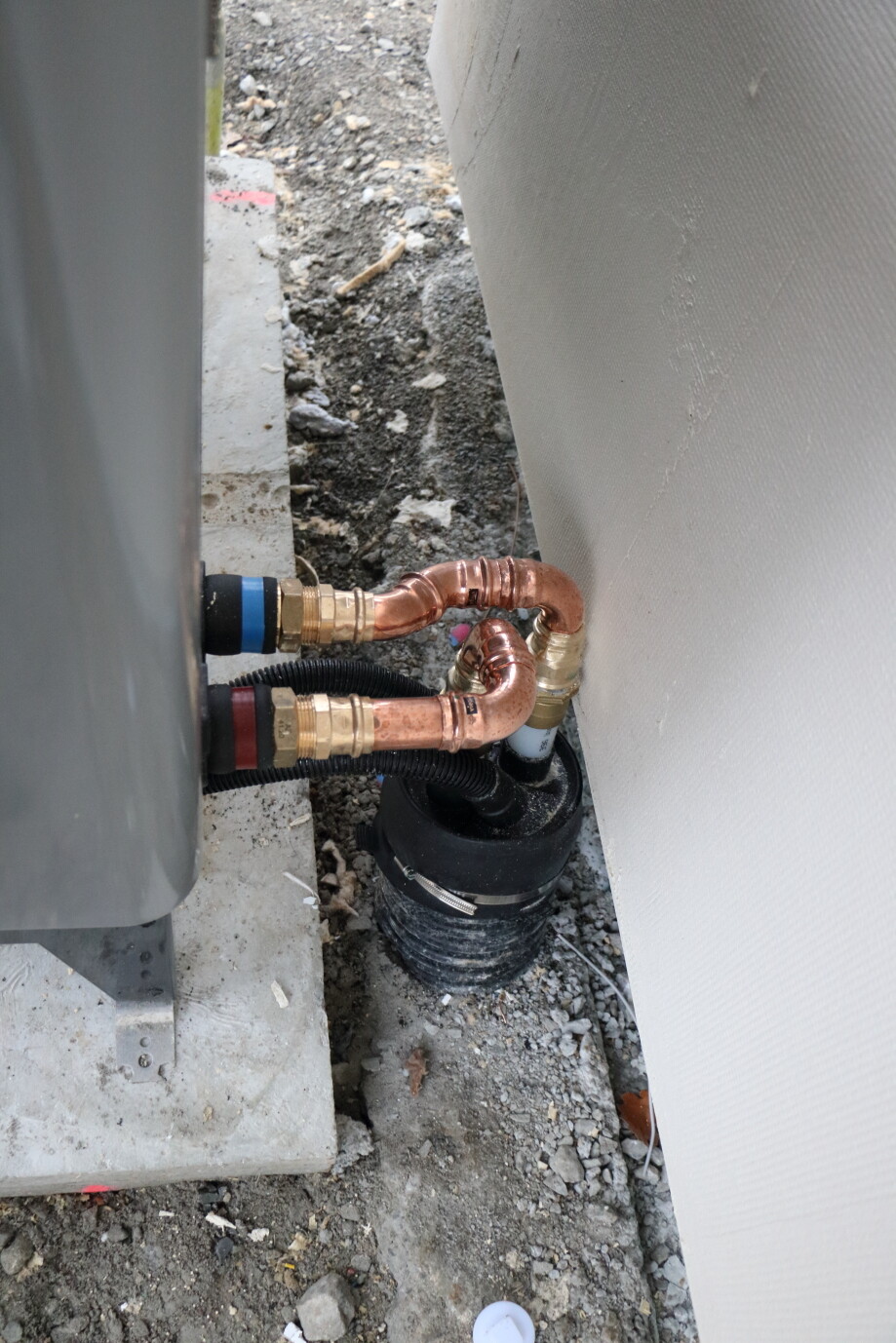 Außen Kupferrohre mit Fittings verbinden sich zu einer schwarzen Pumpe neben einer Wand.
