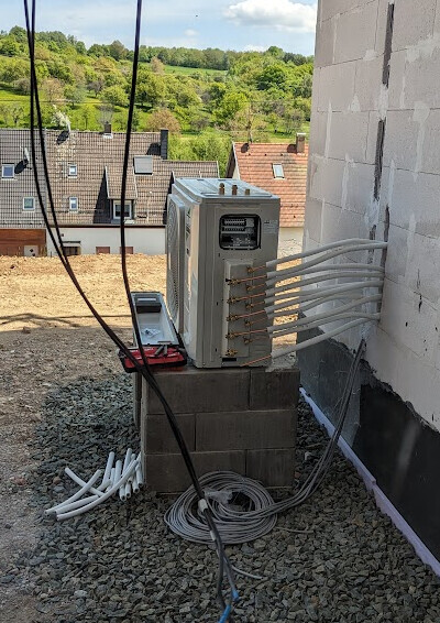 Außen-AC-Einheit auf Betonsteinen, viele Kabel/Rohre, Baugebiet und grüne Hügel im Hintergrund