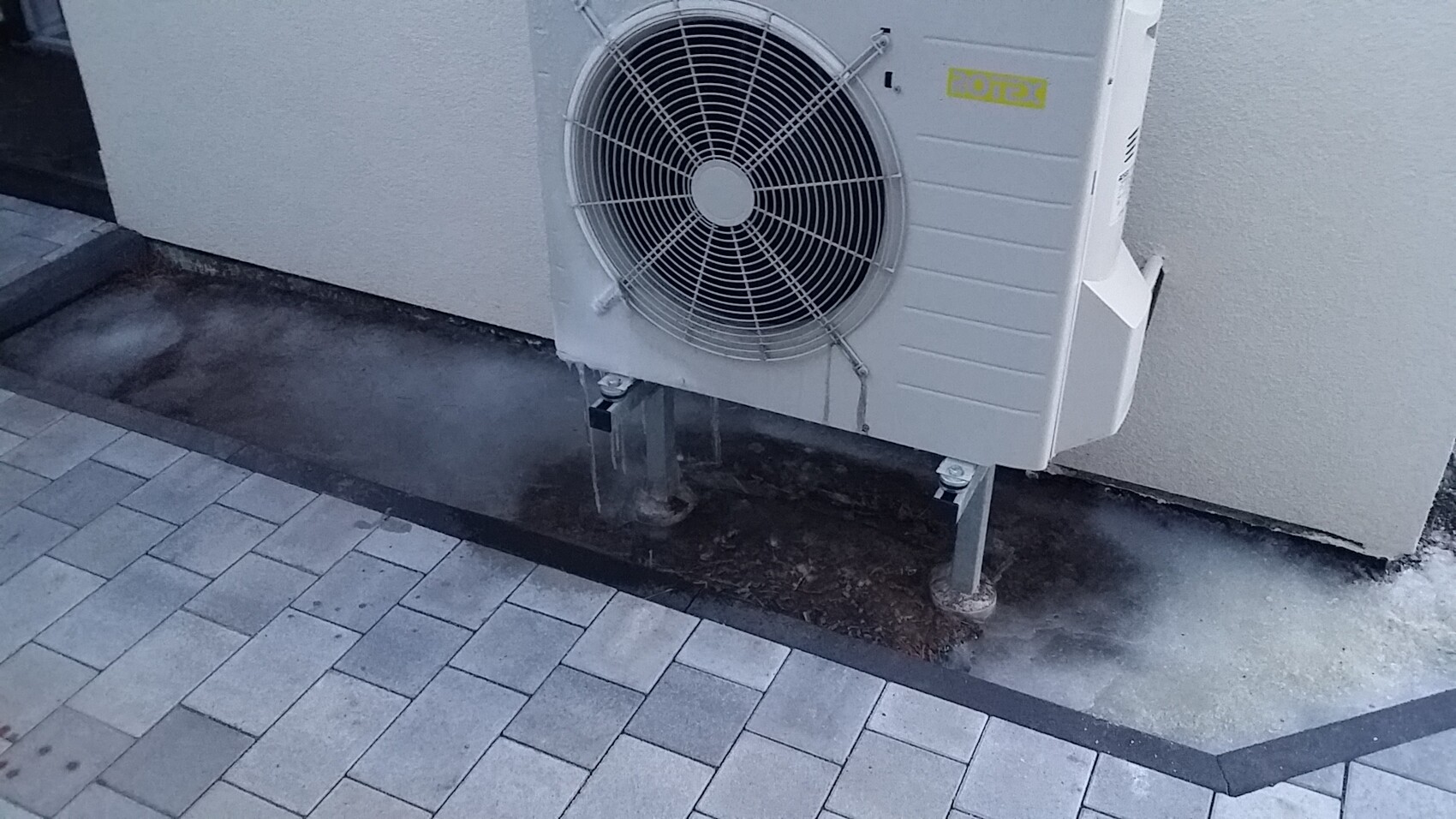 Außengerät einer Klimaanlage mit Lüftergitter, an der Außenwand montiert, auf Stützfüßen.