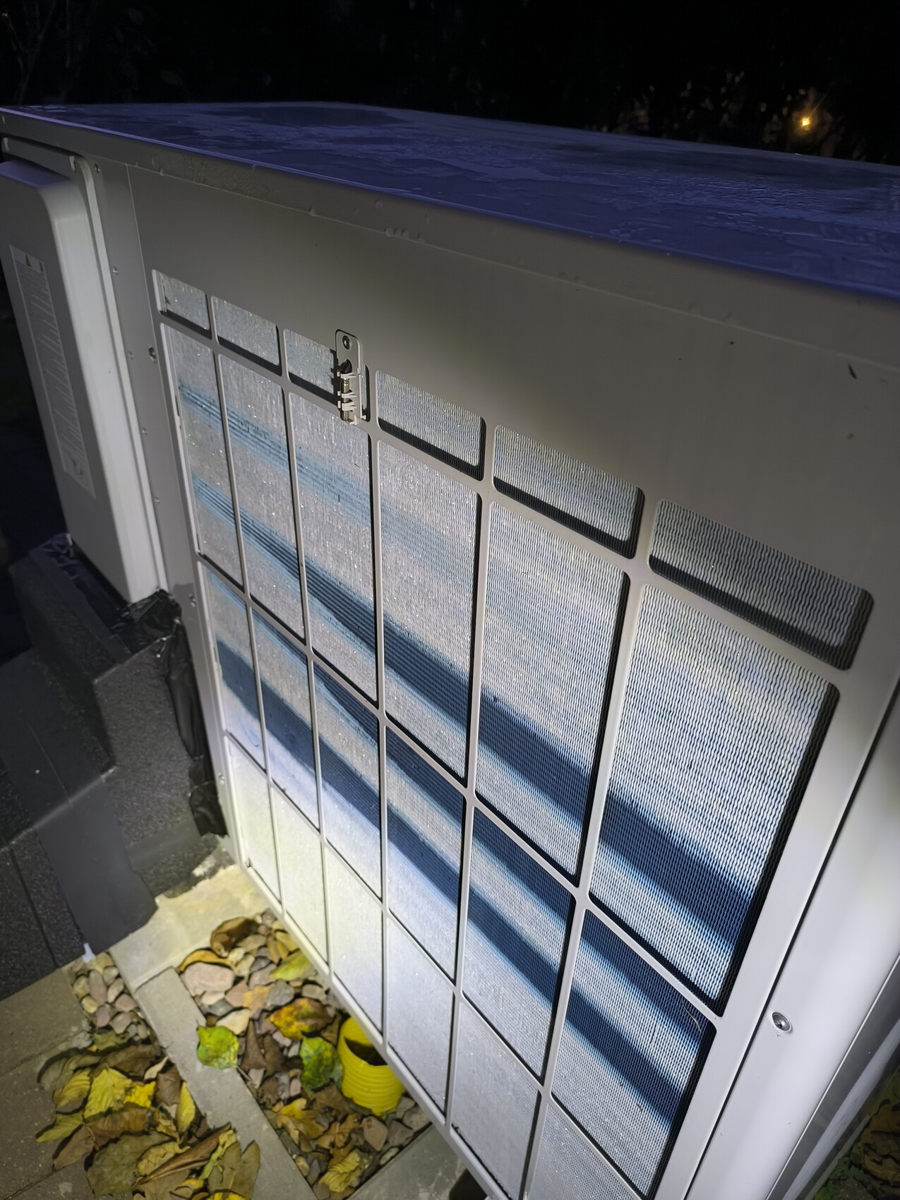 Außengerät einer Klimaanlage mit Gittern im Außenbereich bei Nacht, Blätter am Boden.
