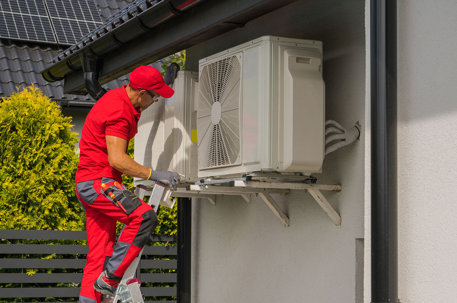 Monteur in rotem Overall auf Leiter repariert Außeneinheit einer Klimaanlage am Haus.
