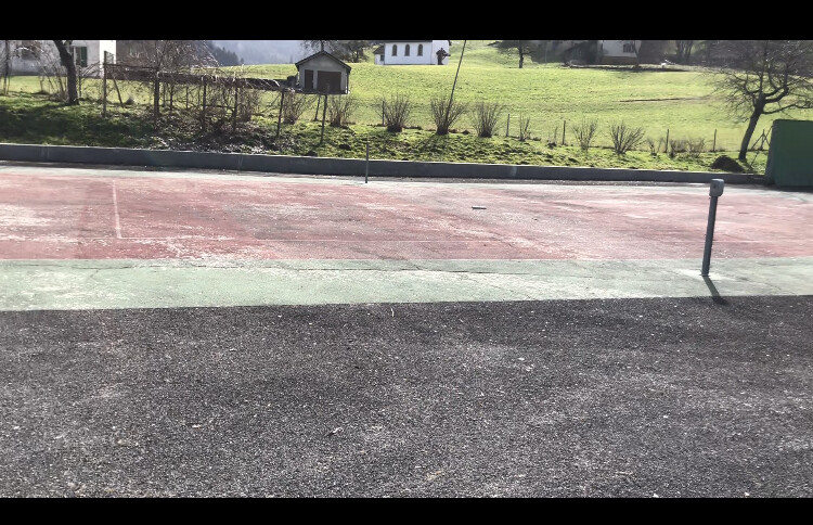 Außenbereich mit rotem Court, grünem Streifen, Asphaltweg, Grasfläche und Häusern im Hintergrund.