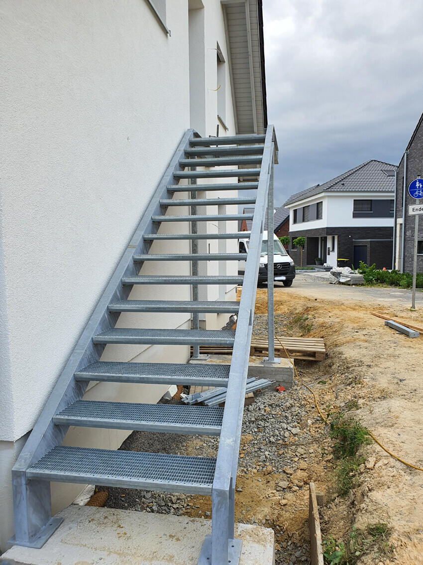 Außenstahltreppe an einem weißen Haus; Baufläche und Wohngebiet im Hintergrund.