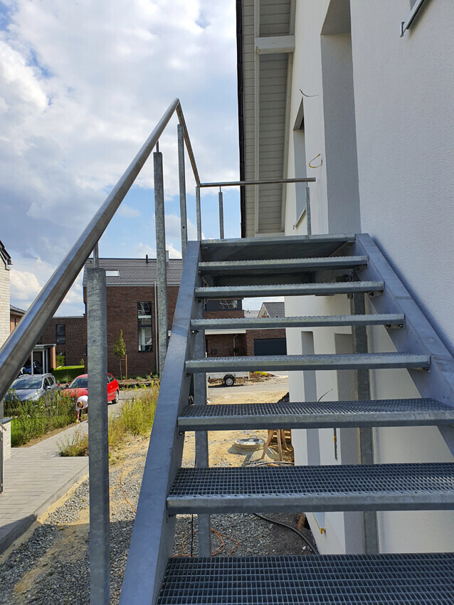 Außentreppe aus Metall an weißer Gebäudefassade, Stufen führen nach oben, Baustellenumgebung im Hintergrund.