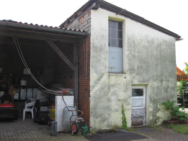 Außenansicht eines alten zweistöckigen Gebäudes mit Carport und Werkstatt im Vordergrund.