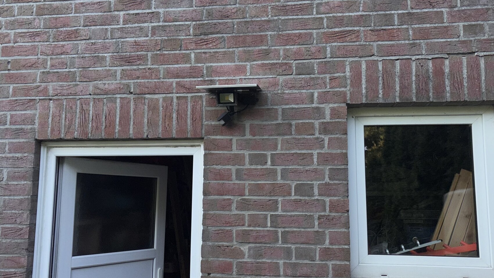 Backsteinfassade mit offener Tür links und Fenster rechts; Außenleuchte über dem Türbereich.