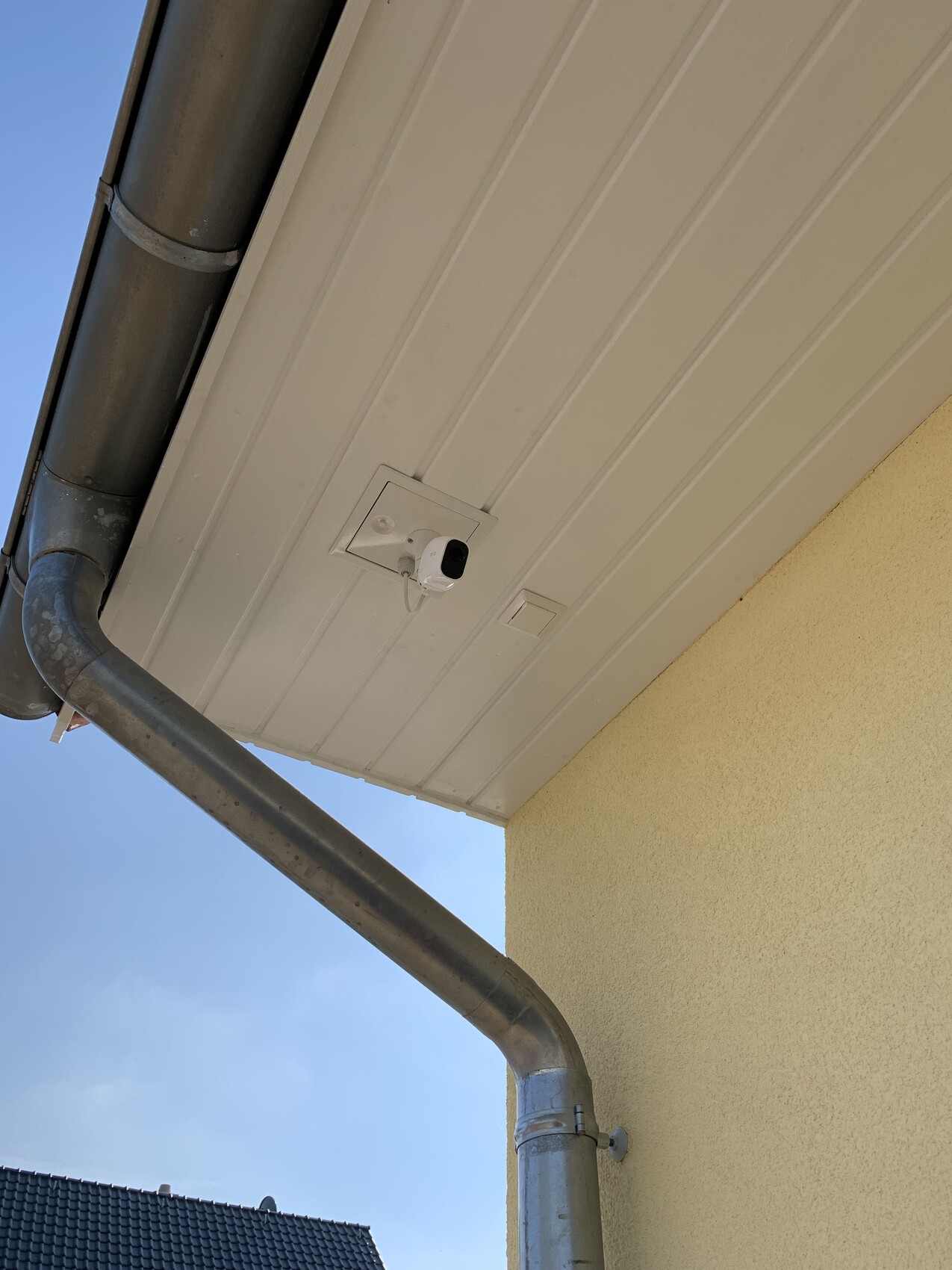 Weiße Außenkamera unter Dachverkleidung neben einer Metall-Regenrinne an gelber Hauswand.