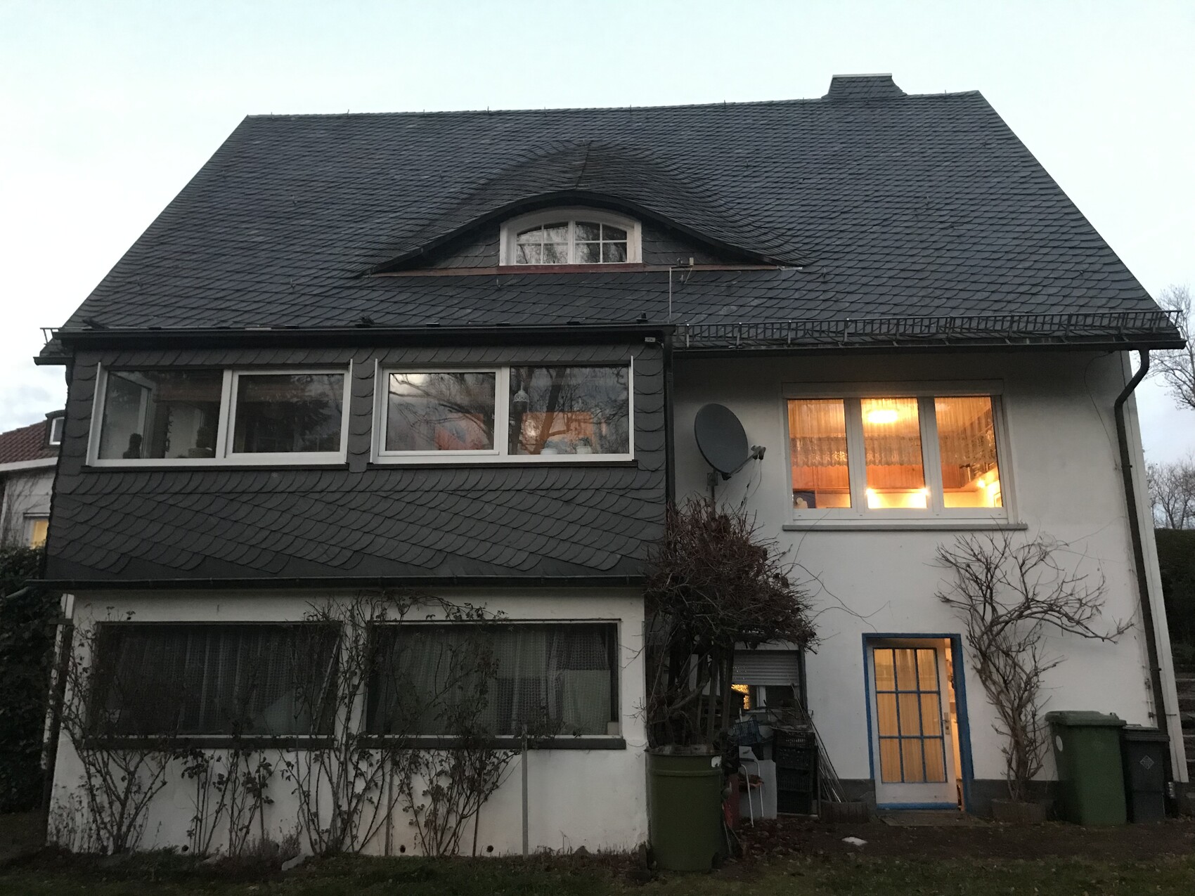 Zweistöckiges Haus mit dunklem Schieferdach, Dachgaube, Fensterfront und Satellitenschüssel.