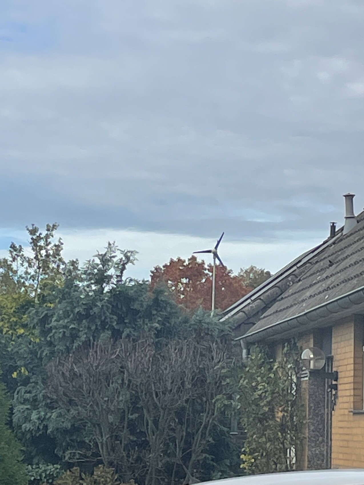 Außenansicht eines Hauses mit geneigtem Dach, Windrad im Hintergrund und Bäumen