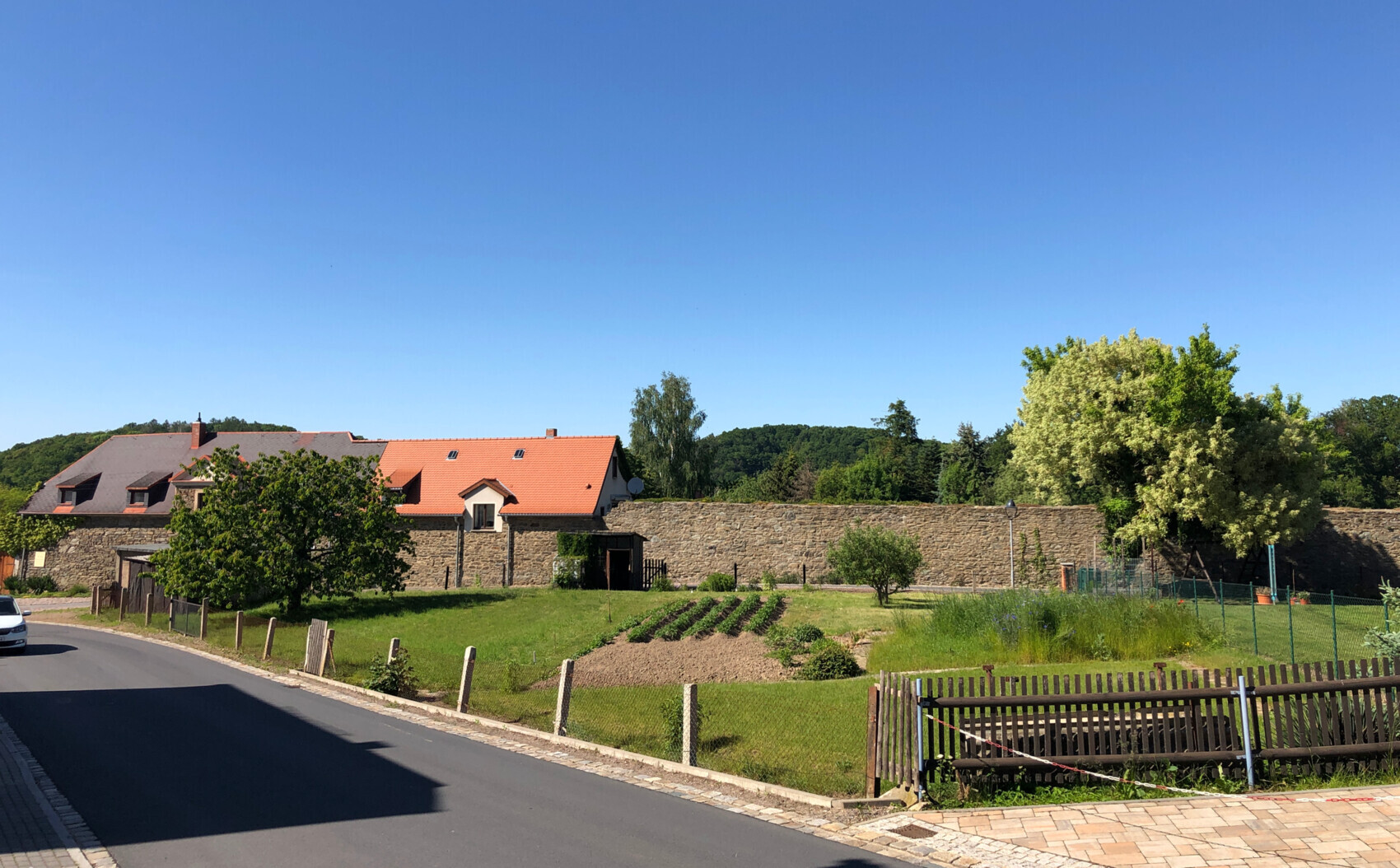 Ländliche Szene: Steinmauer, Häuser mit roten Ziegeldächern, Gartenbeete und Bäume vor Grünfläche.