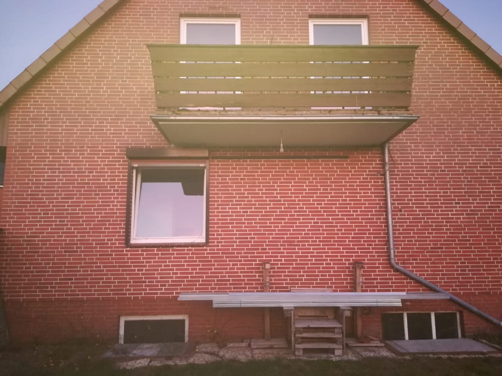 Rotes Ziegelhaus-Rückseite mit Balkon, Fenster, Fallrohr und Paletten vor dem Haus.