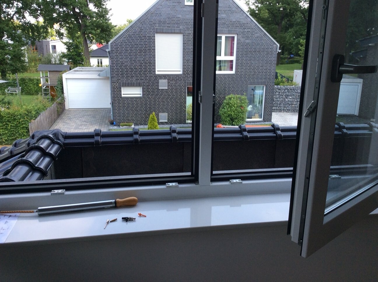 Blick aus geöffnetem Fenster auf ein graues Backsteinhaus mit Garage und Garten
