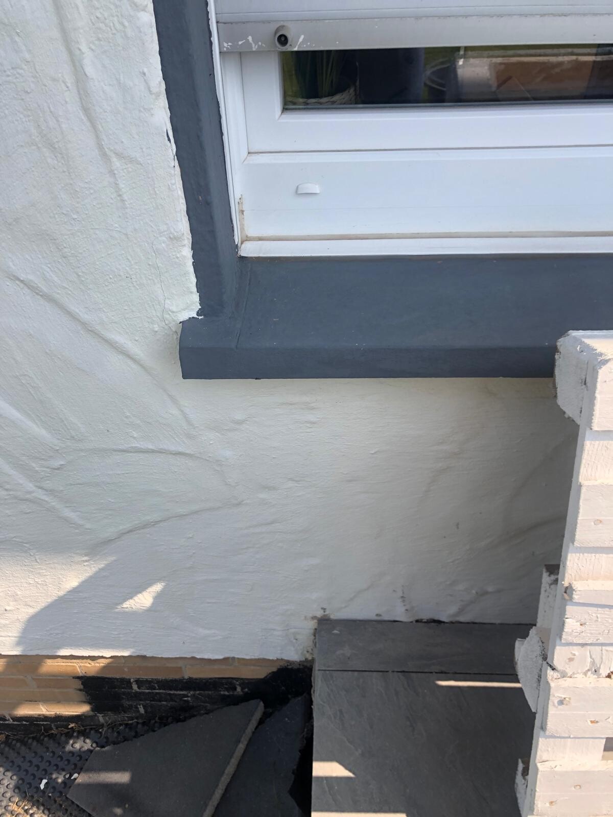 Außenfenster mit weißem Rahmen und grauer Fensterbank an weißer Wand, darunter dunkle Stufen.