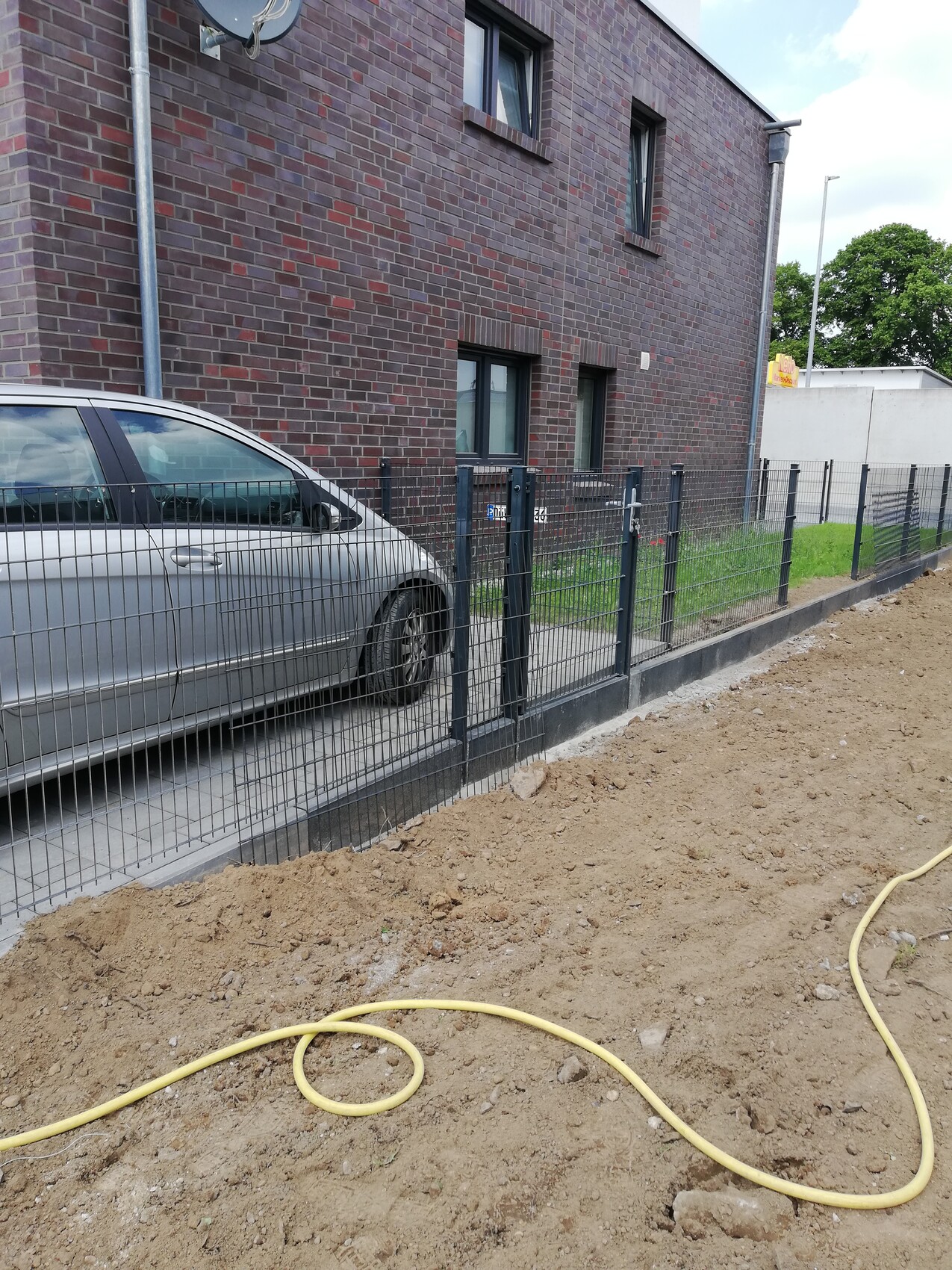 Geparktes silbernes Auto neben Metallzaun vor rotem Backsteinhaus; gelbe Schlauch liegt auf Baugrund.