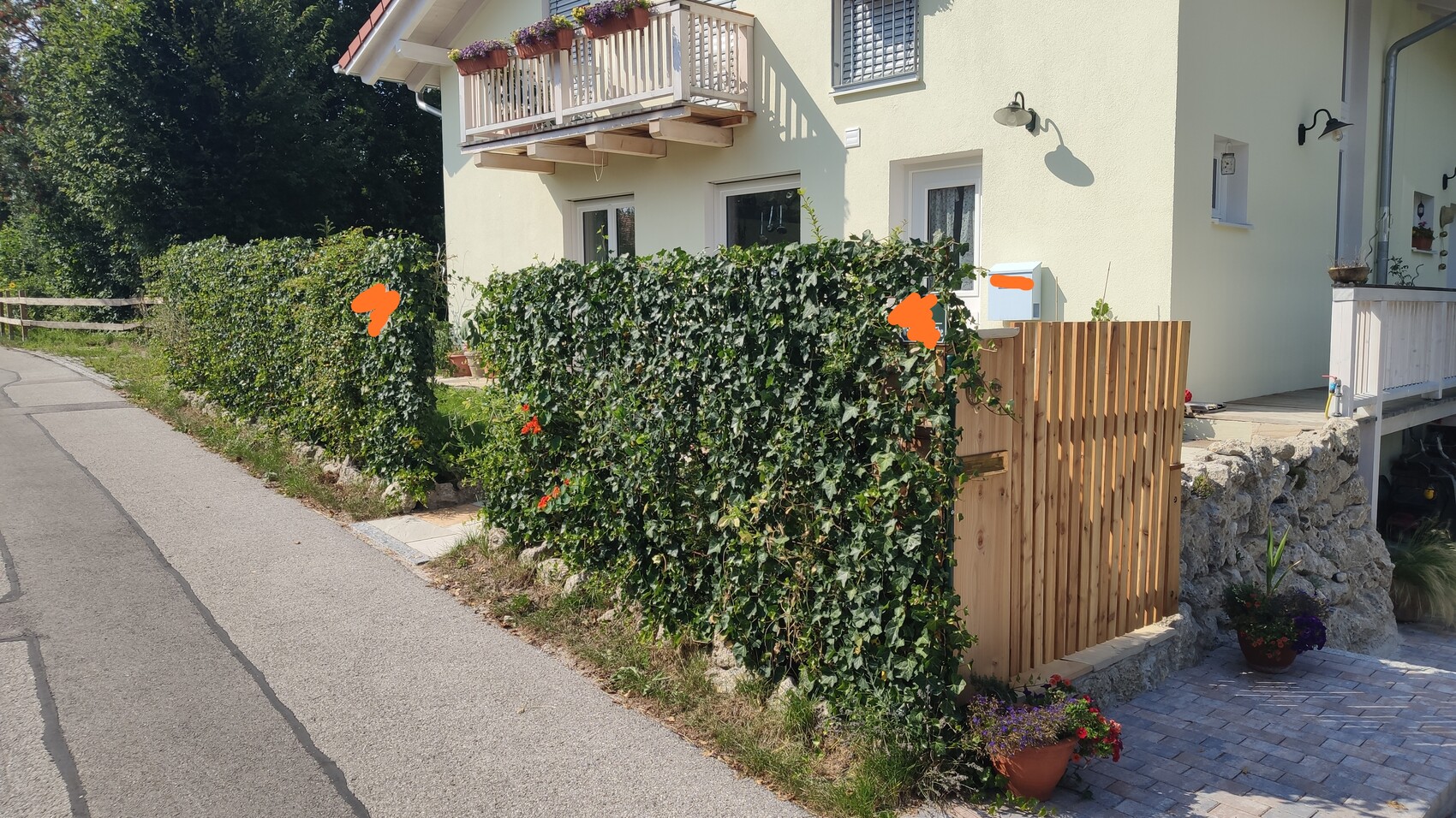 Gelbes Haus mit Balkon, grüne Hecke als Sichtschutz, Holztor und Pflanzkübel an Steinmauer