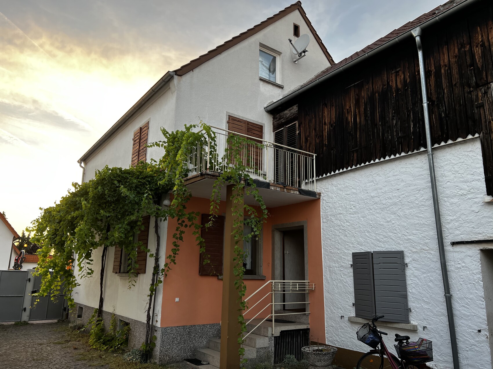 Zweistöckiges Haus mit Balkon, Kletterpflanzen, braune Fensterläden und Fahrrad davor.