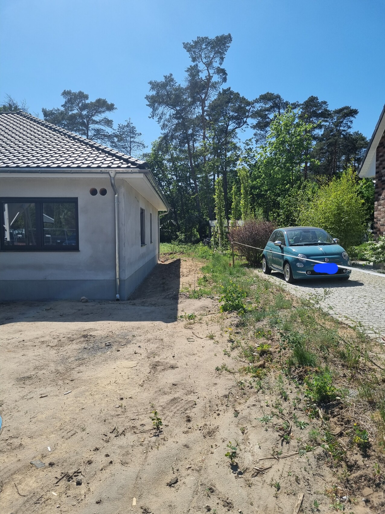 Ein Haus links, unbefestigter Hof vorne, blau-grünes Auto auf gepflasterter Einfahrt, Wald dahinter.