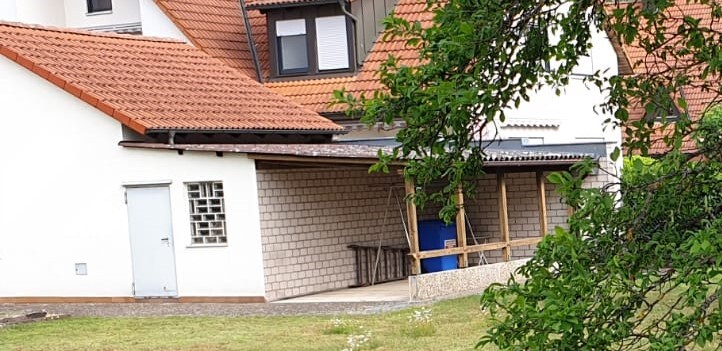 Weißes Haus mit rotem Ziegeldach; überdachte Veranda aus Holzpfosten, blaue Fässer, Baum.