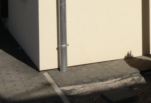 Beige Gebäudewand mit senkrechtem Regenrohr, gepflastertem Gehweg, Bordstein und Schatten.