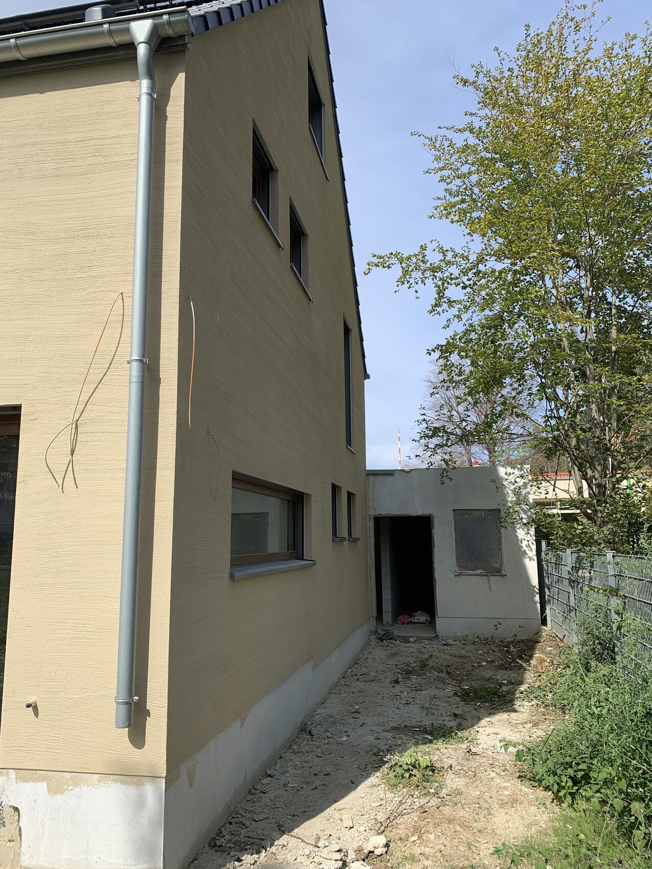 Seitliche beige Hauswand mit grauer Regenrinne, Fensterfront, Nebengebäude und Gartenweg.