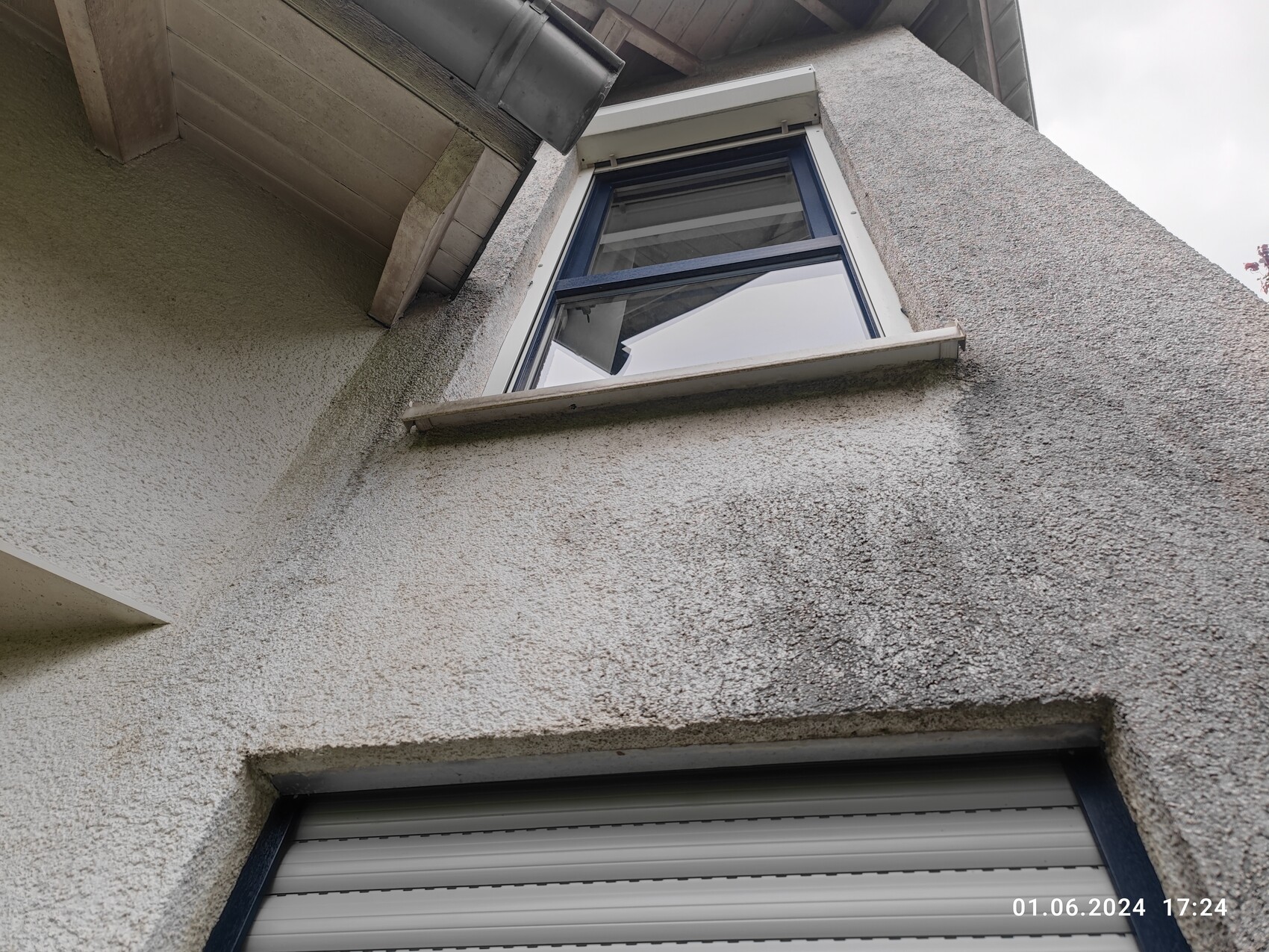 Außenwand eines Hauses mit grobem Putz, oben Fenster mit gebrochener Scheibe, unten Rollladen.