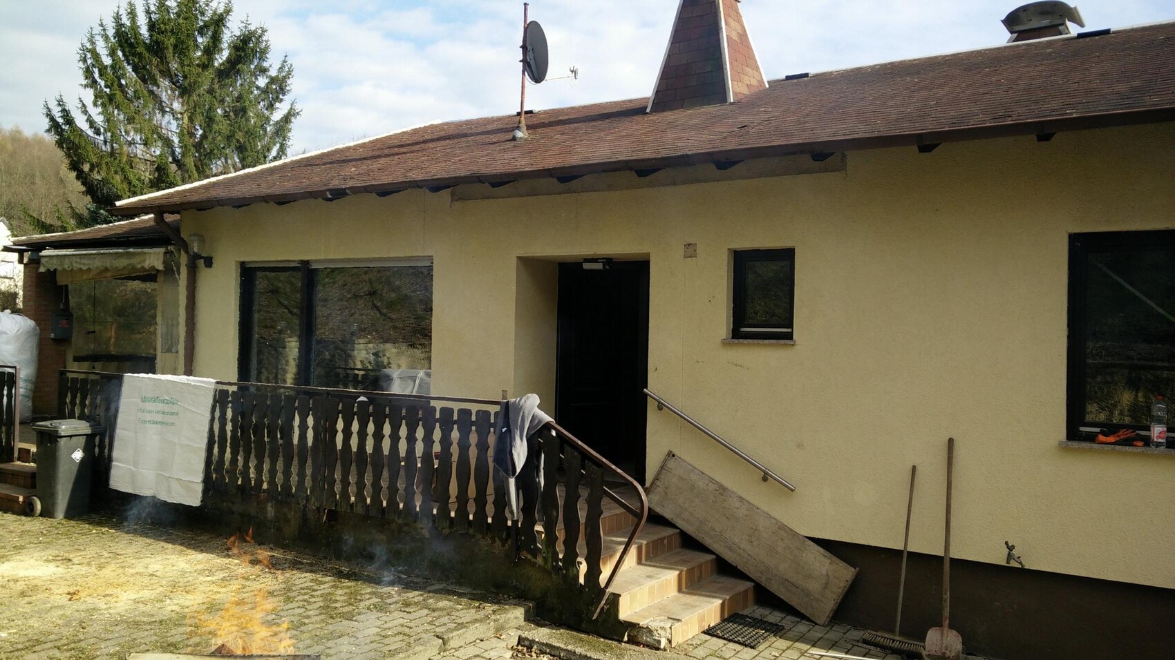 Außenansicht eines einstöckigen gelben Hauses mit Eingangstür, Geländer und Terrasse.