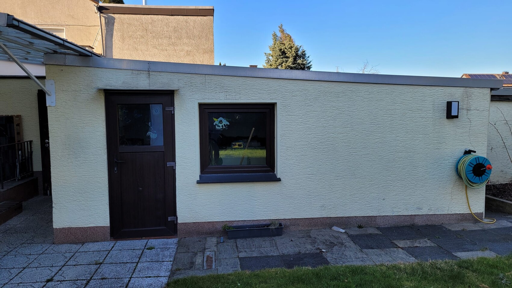Kleines Anbaugebäude mit heller Wand, dunkler Tür und Fenster; Schlauchhalter an der Wand.