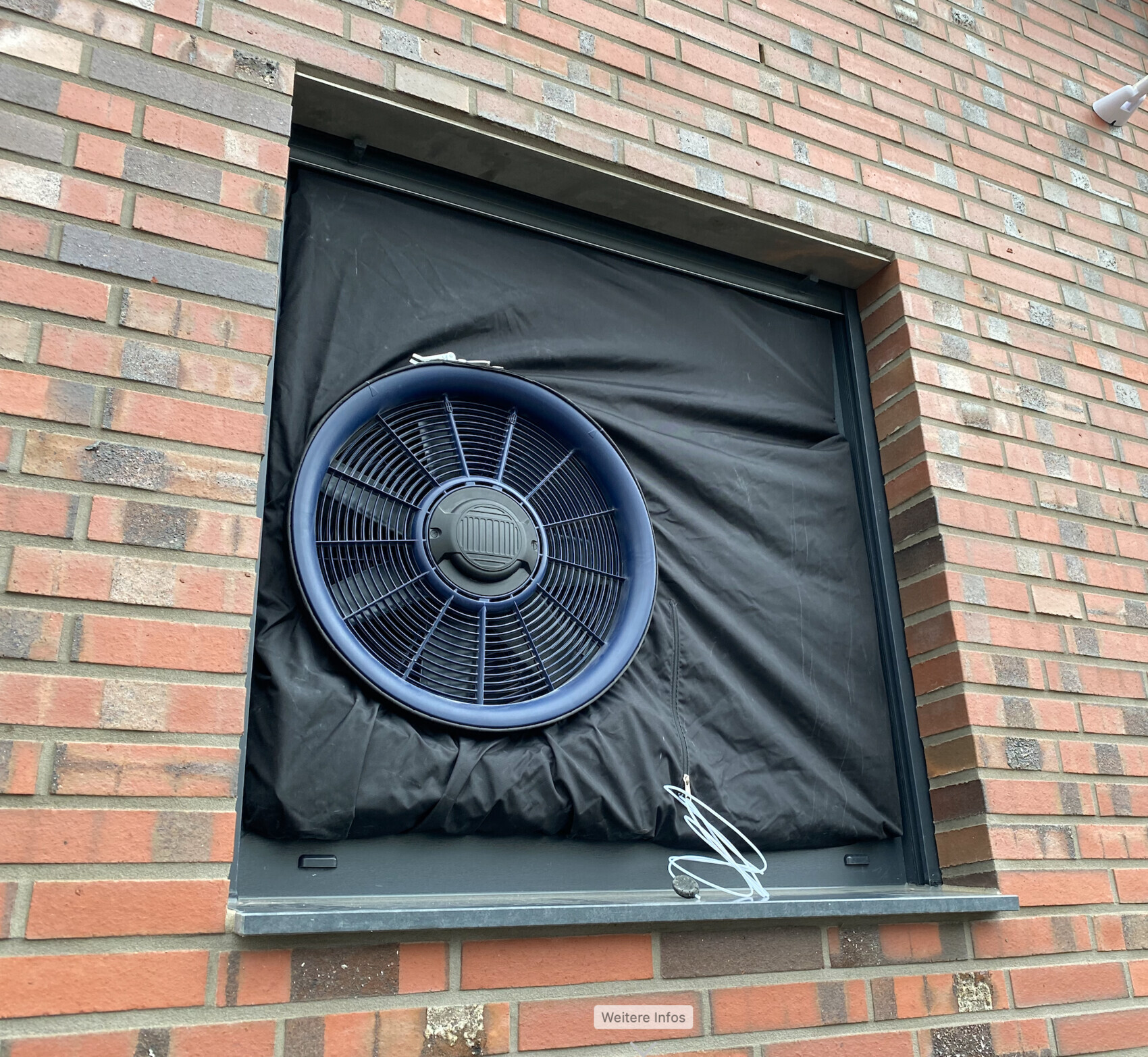 Großer runder blauer Ventilator hinter schwarzer Abdeckung in einer Backstein-Fensteröffnung.