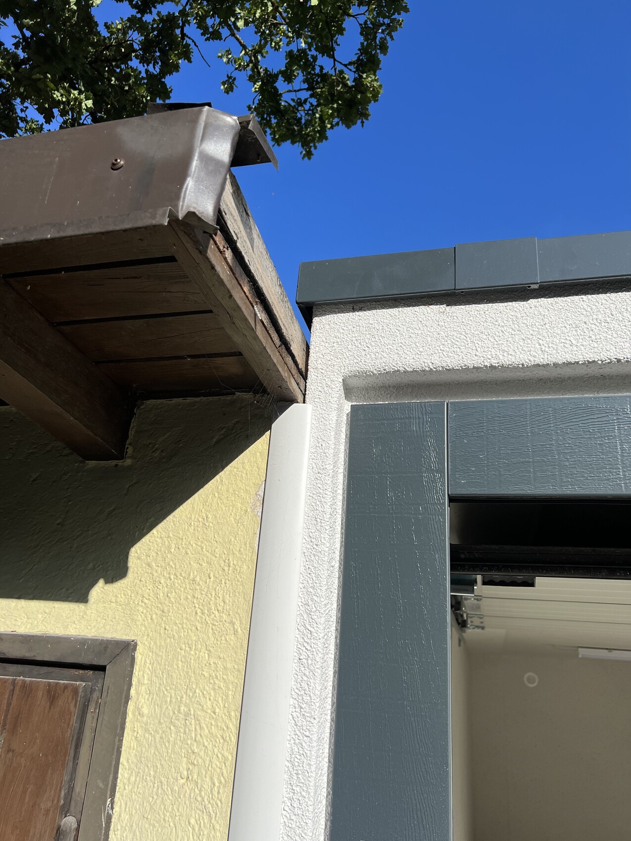Außenansicht: Dachüberstand aus Holz, gelbe Wand, graue Türrahmen, blauer Himmel.