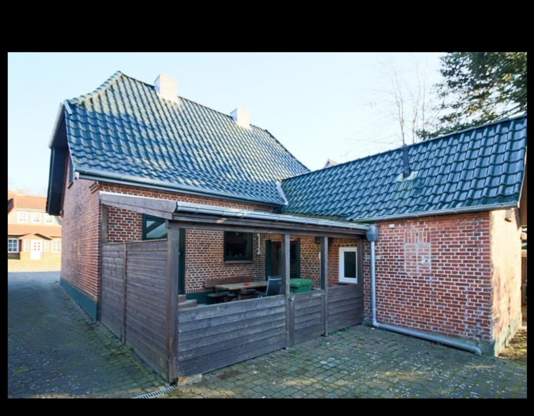 Ein L-förmiges Backsteinhaus mit dunkelgrauen Dachziegeln, Holzwand-Veranda und gepflastertem Hof.