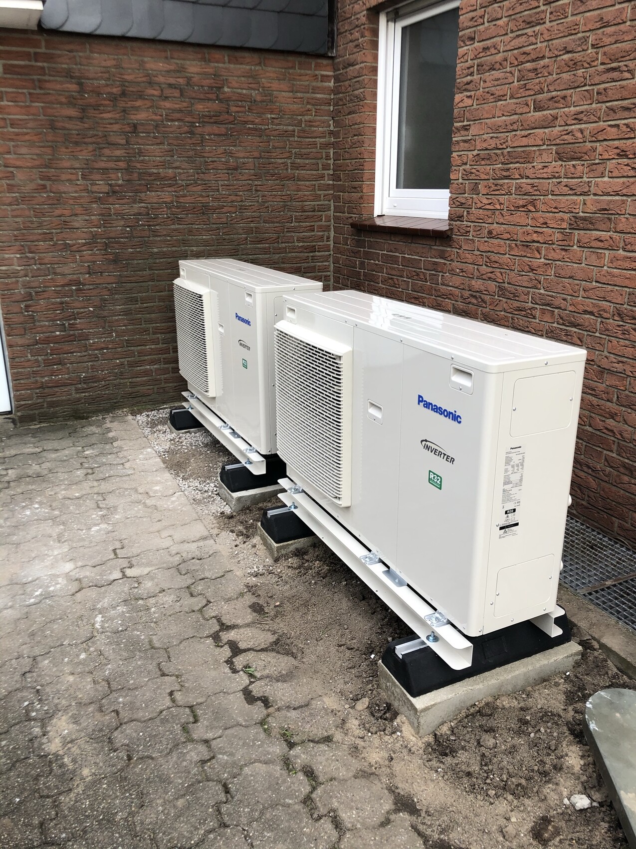 Zwei Panasonic-Außengeräte einer Klimaanlage vor roter Backsteinwand, auf Betonfundamenten montiert.