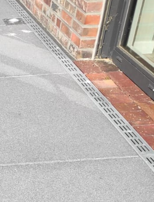 Gehweg mit Metallrinne entlang einer roten Backsteinwand neben einer Glasfassade eines Geschäfts.