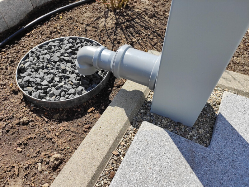 Außeninstallation: PVC-Rohrbogen am Metallpfosten, Kiesring daneben und Betonstufen.