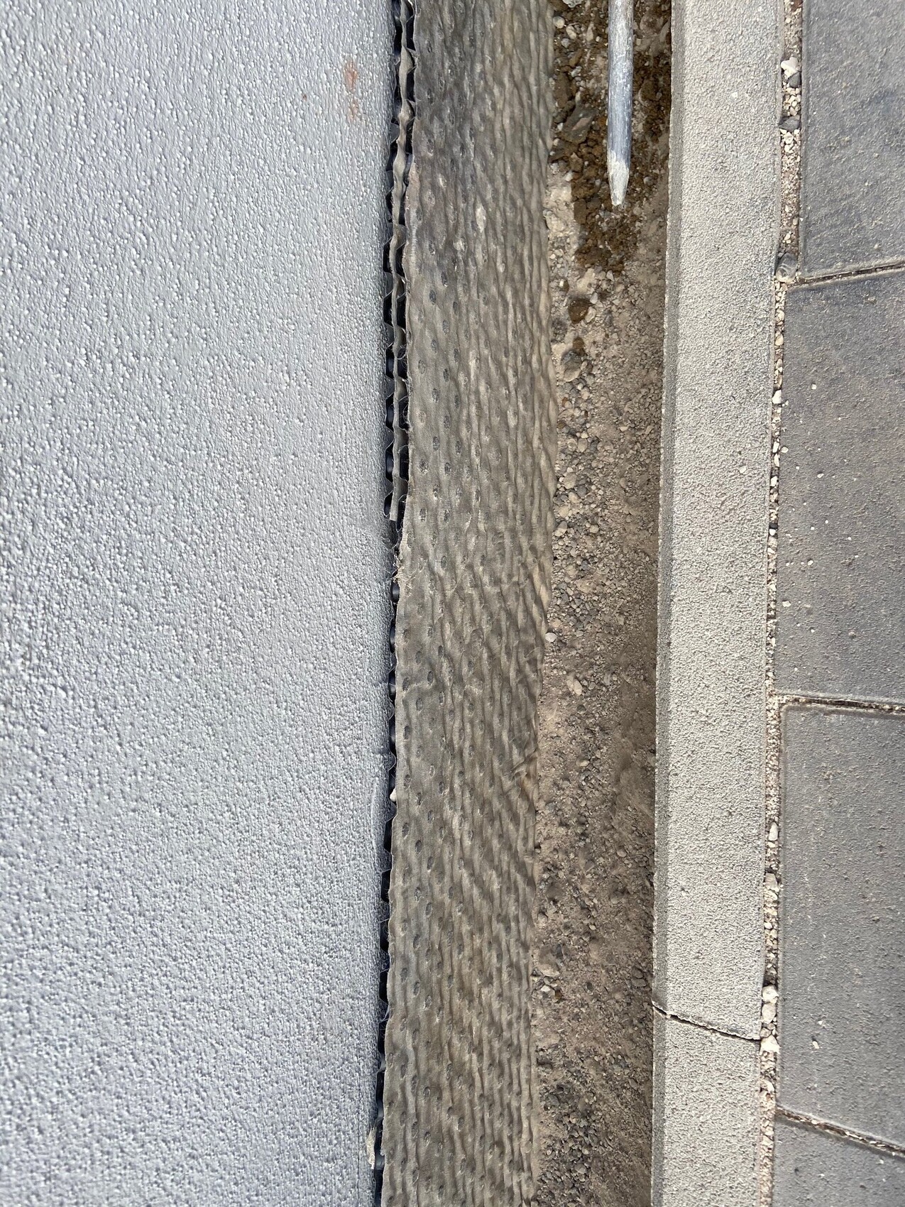 Spalt zwischen grauer Wand und Pflasterkante mit dunklem, geriffeltem Profil und Erdreich daneben.