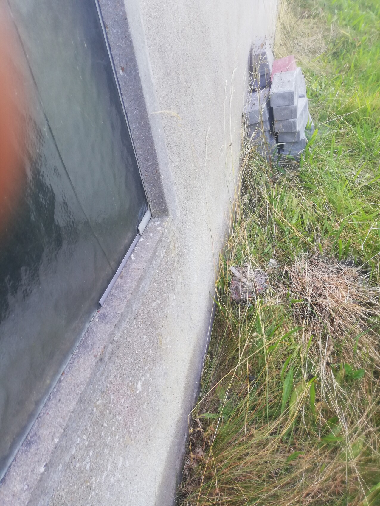 Außenwand aus Beton mit Glastür, daneben gestapelte Betonblöcke und Gras.